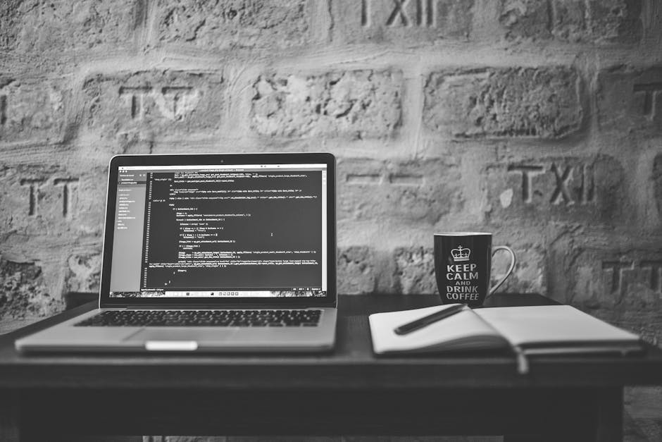A black-and-white photograph of a workspace with a laptop displaying lines of coding on the screen, placed on a dark wooden desk against a textured brick wall. To the right of the laptop is an open no